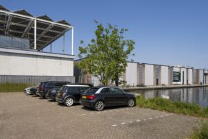 Parkeerplaats aan de Anthony Fokkerweg met geparkeerde auto's naast een gracht en industrieel gebouw met te huur-bord.